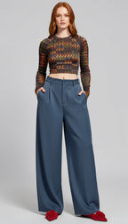 A model stands against a light gray studio background wearing a long-sleeve cropped knit top with a brown, orange, and black chevron pattern, paired with high-waisted slate blue wide-leg trousers and red pointed flats.
