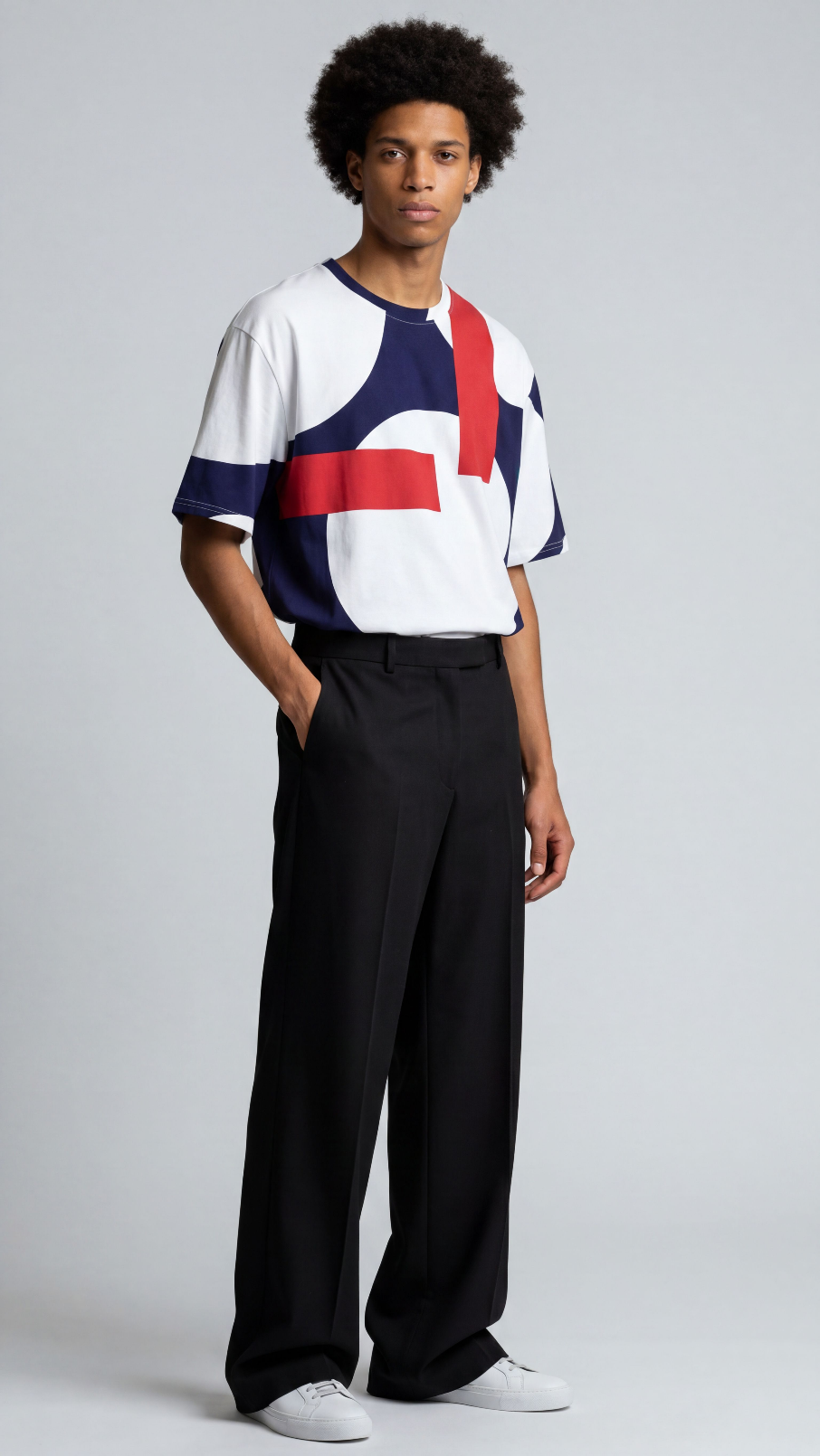 A man modeling a short-sleeve crewneck t-shirt with a bold geometric color-block pattern in white, navy, and red, paired with black wide-leg trousers and white sneakers, against a light gray studio background.