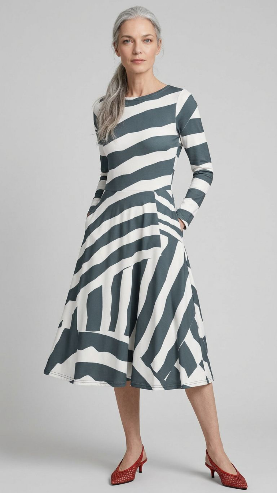 A woman modeling a long-sleeve midi dress with a bold horizontal stripe pattern in dark teal and off-white. She is wearing red pointed-toe heels against a plain light gray studio background.