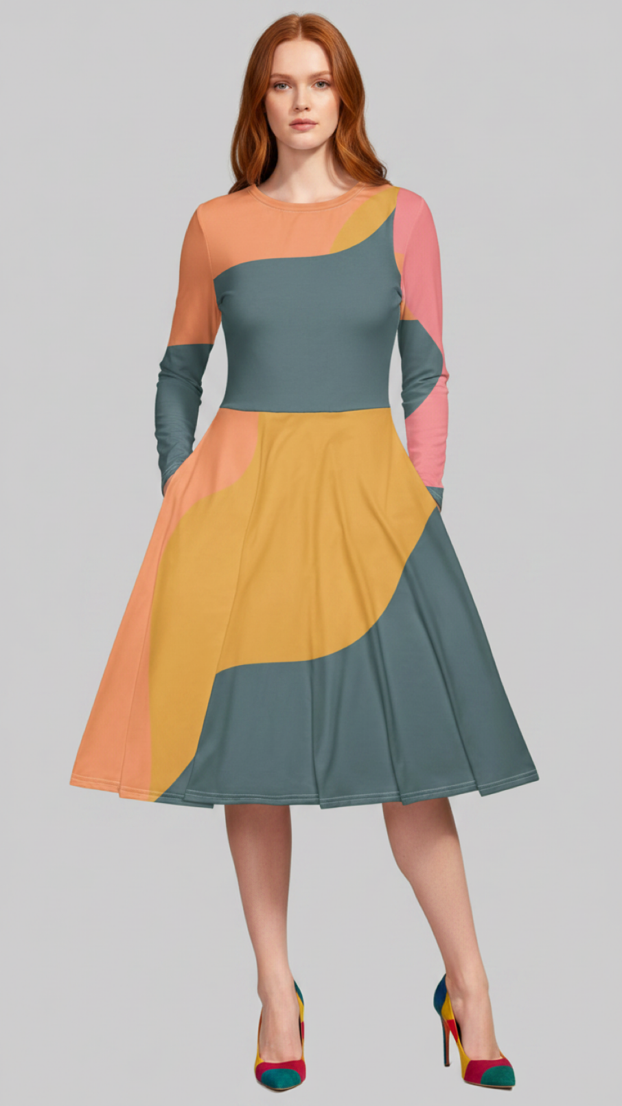 A woman modeling a color-block midi dress with long sleeves, featuring a teal blue bodice, peach and pink upper sleeves, a mustard yellow skirt, and a teal blue lower skirt panel, paired with multicolor high heels, against a solid gray background.